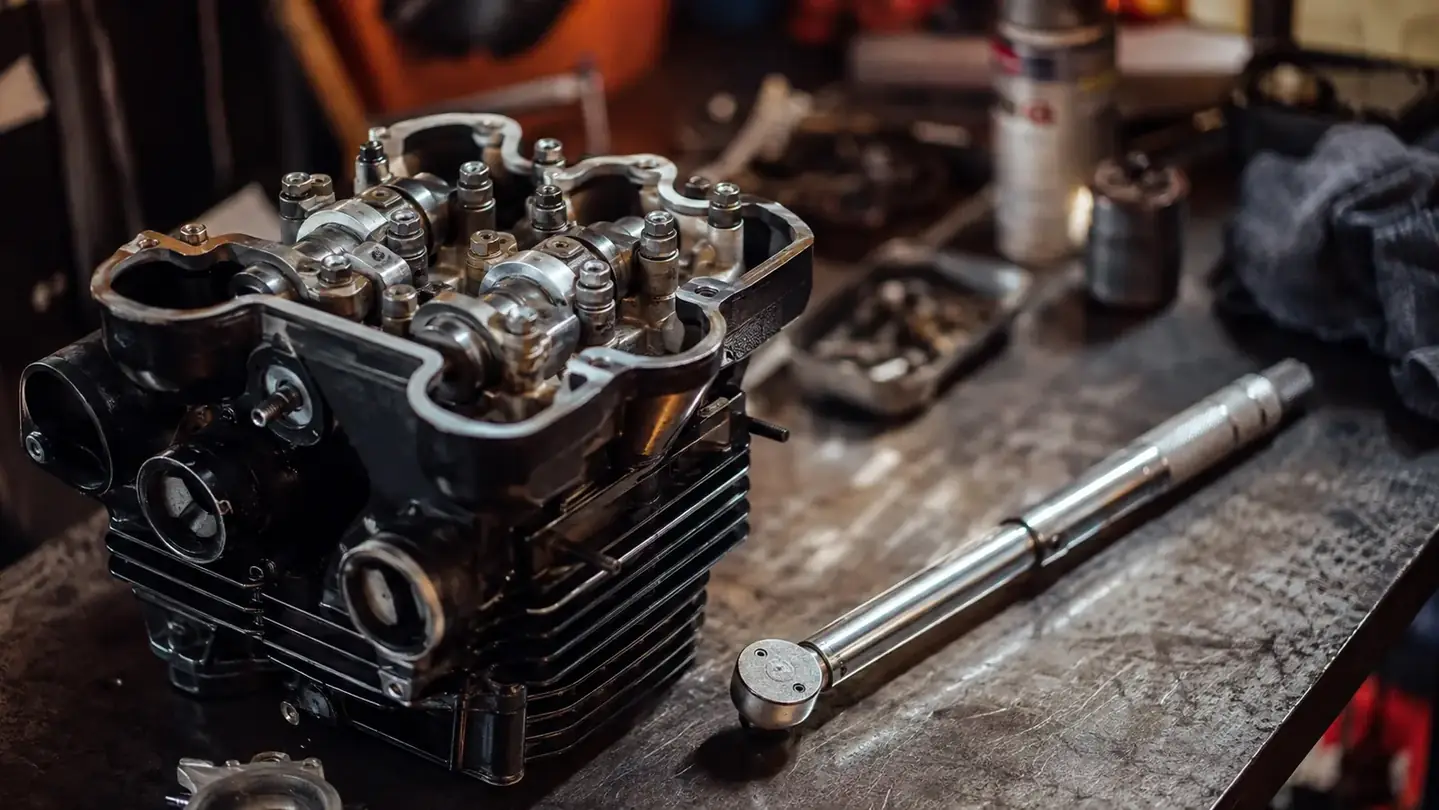 A motorcycle engine sitting on a workbench with its valve cover off, exposed cam caps and head bolts, a click-type torque wrench resting on the bench nearby